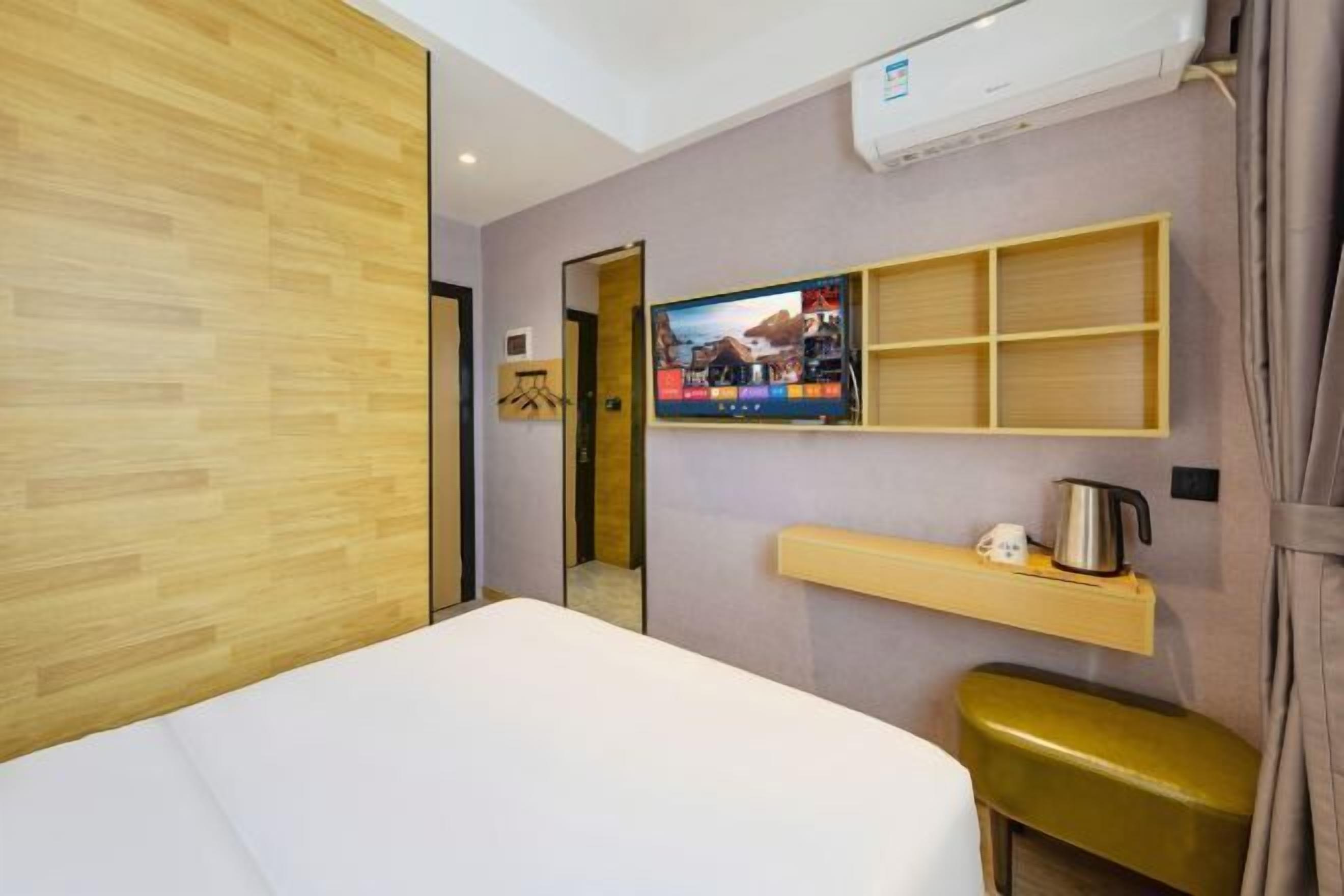 Quarto de hotel com uma cama, televisão fixada na parede, um pequeno armário com chaleira e um banco de madeira.
