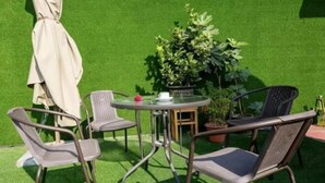 Une terrasse avec une table ronde et quatre chaises, entourée de gazon synthétique et de plantes en pot.