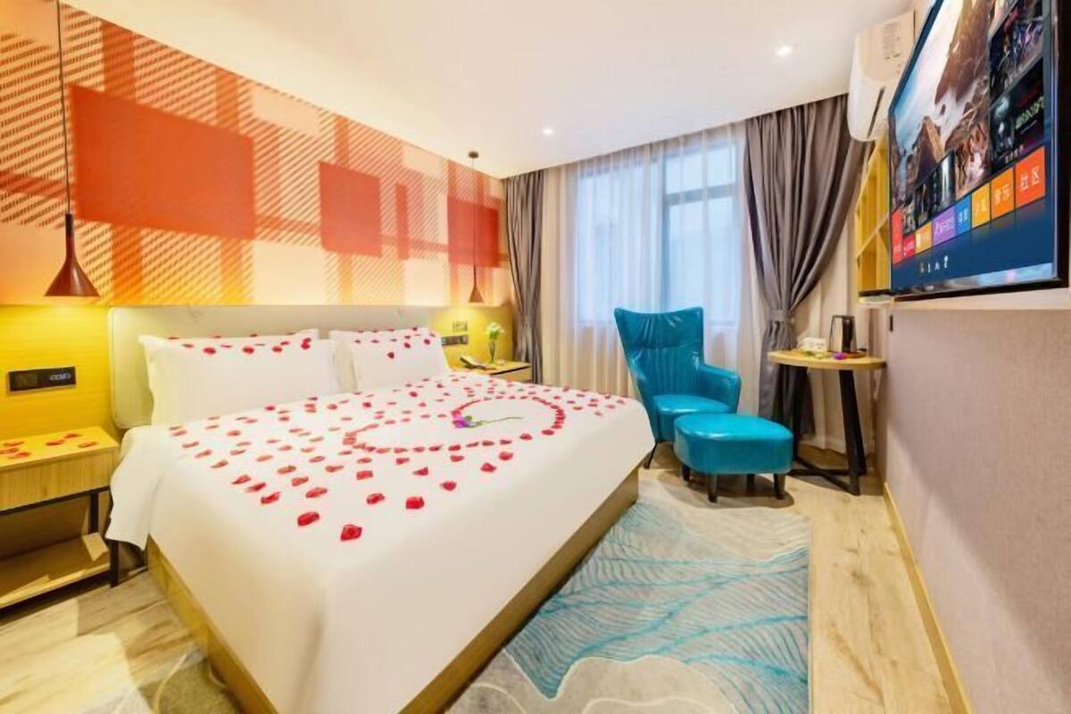Habitación de hotel con una cama decorada con pétalos de rosa, un sillón azul, una mesita y un televisor montado en la pared.