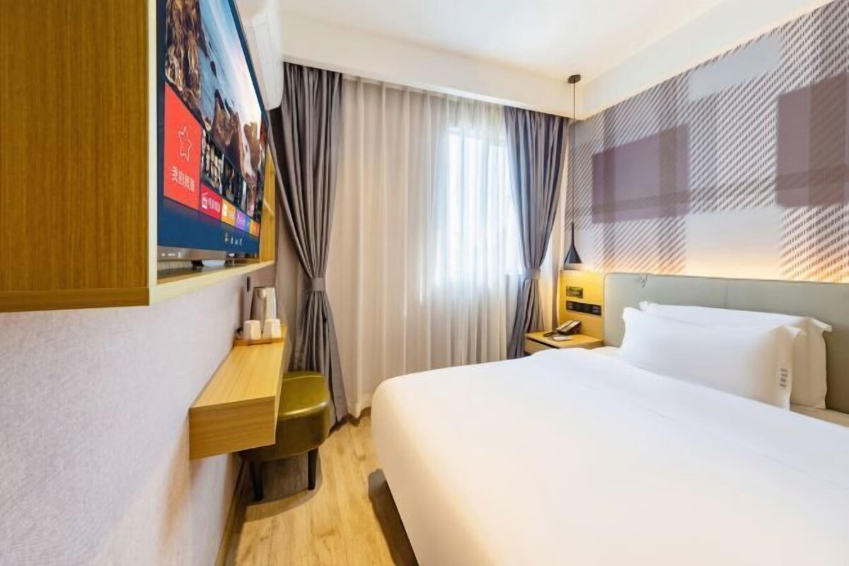 A hotel room with a large bed, a TV on the wall, a desk, and a window with curtains.