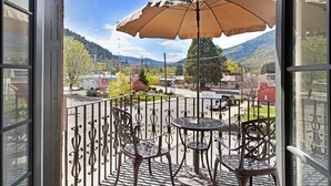 Ein Balkon mit schmiedeeisernen Stühlen und einem Tisch, mit Blick auf eine Straße, an deren Seite Gebäude stehen und im Hintergrund ein Berg zu sehen ist.