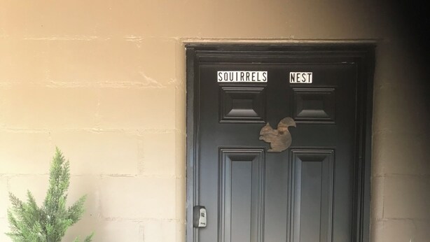 Une porte noire sur laquelle est accrochée une enseigne indiquant « NID D’ÉCUREUILS », un petit pot de fleurs placé à gauche et un paillasson sur lequel est écrit « BIENVENUE ».