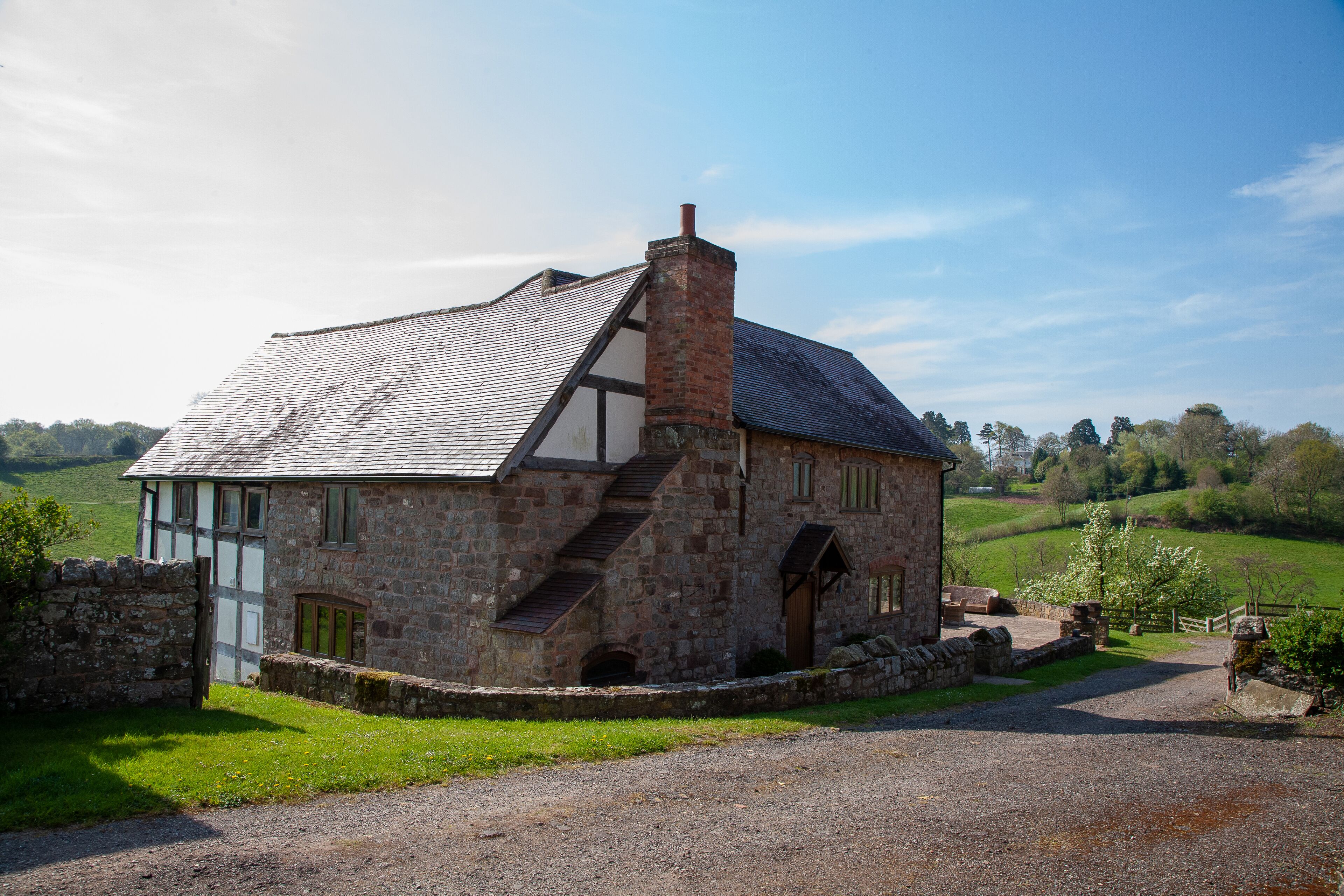 A stone house with a sloped roof and a chimney, surrounded by a gravel driveway and a stone wall.