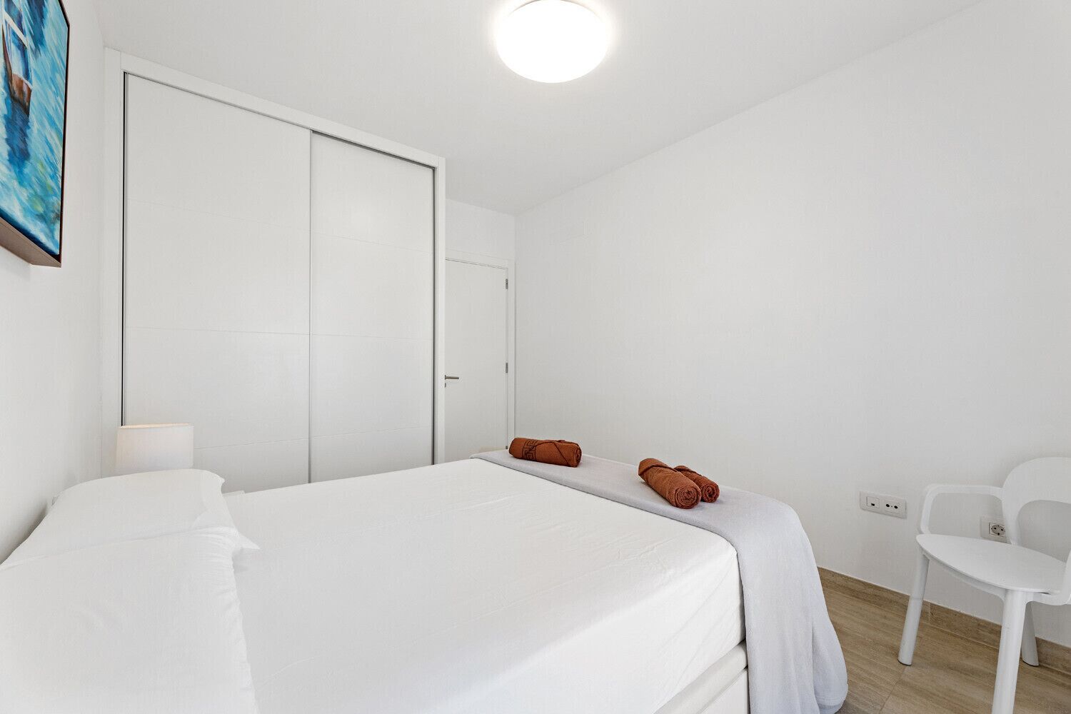 A bedroom with a bed, a white wardrobe, a chair, and a painting on the wall.