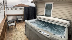 A hot tub on a wooden deck with a white fence, a window, and a chair. -  The Bear Cave w hot tub in the quaint village of Aurora (Hardin)