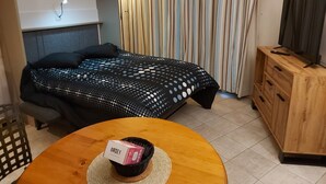 Una habitación ordenada con una cama, un televisor sobre un mueble de madera y una mesa con una cesta que contiene una caja etiquetada “ORSET”.