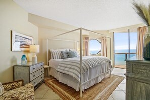 Una cama con dosel y vista al mar, una mesita de noche con lámpara, y un aparador de mimbre.