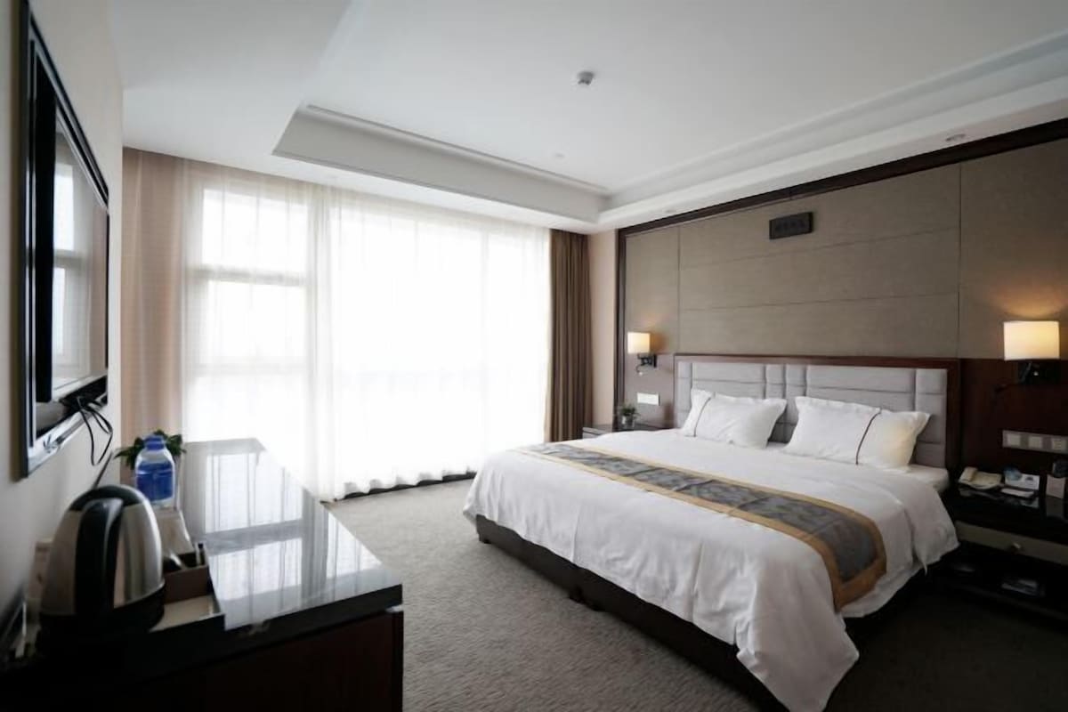 A hotel room with a large bed, a desk with a kettle, a TV, and a window with curtains.