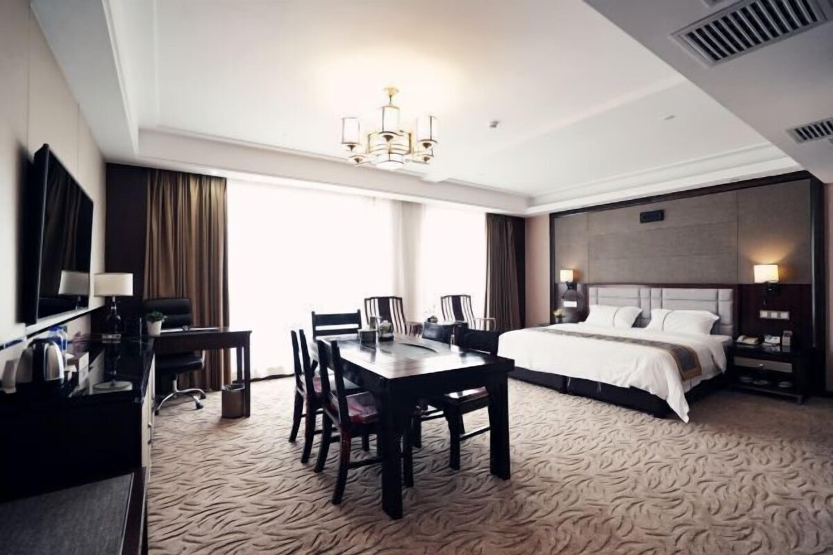 A hotel room with a bed, a desk, a chair, a table, a chandelier, and a large window.