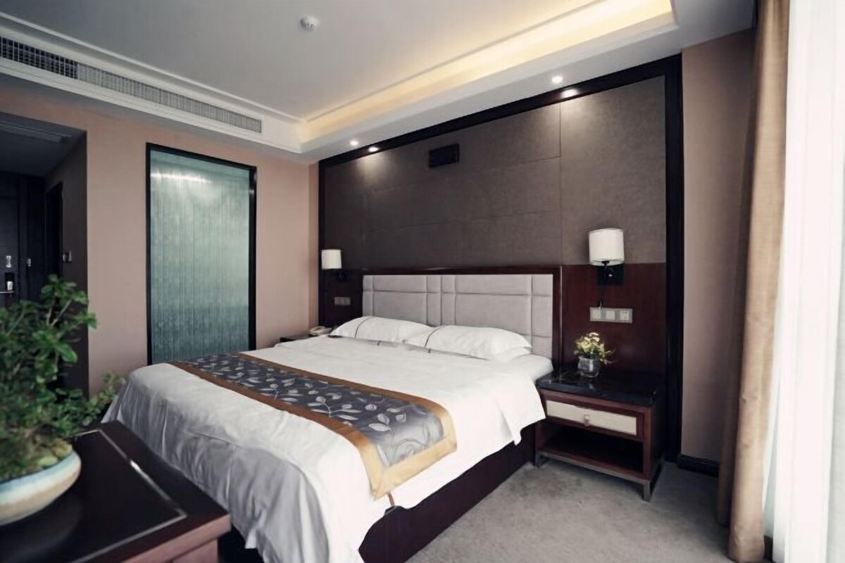A hotel room with a large bed, bedside tables, a nightstand, a potted plant, and a view of the hallway.