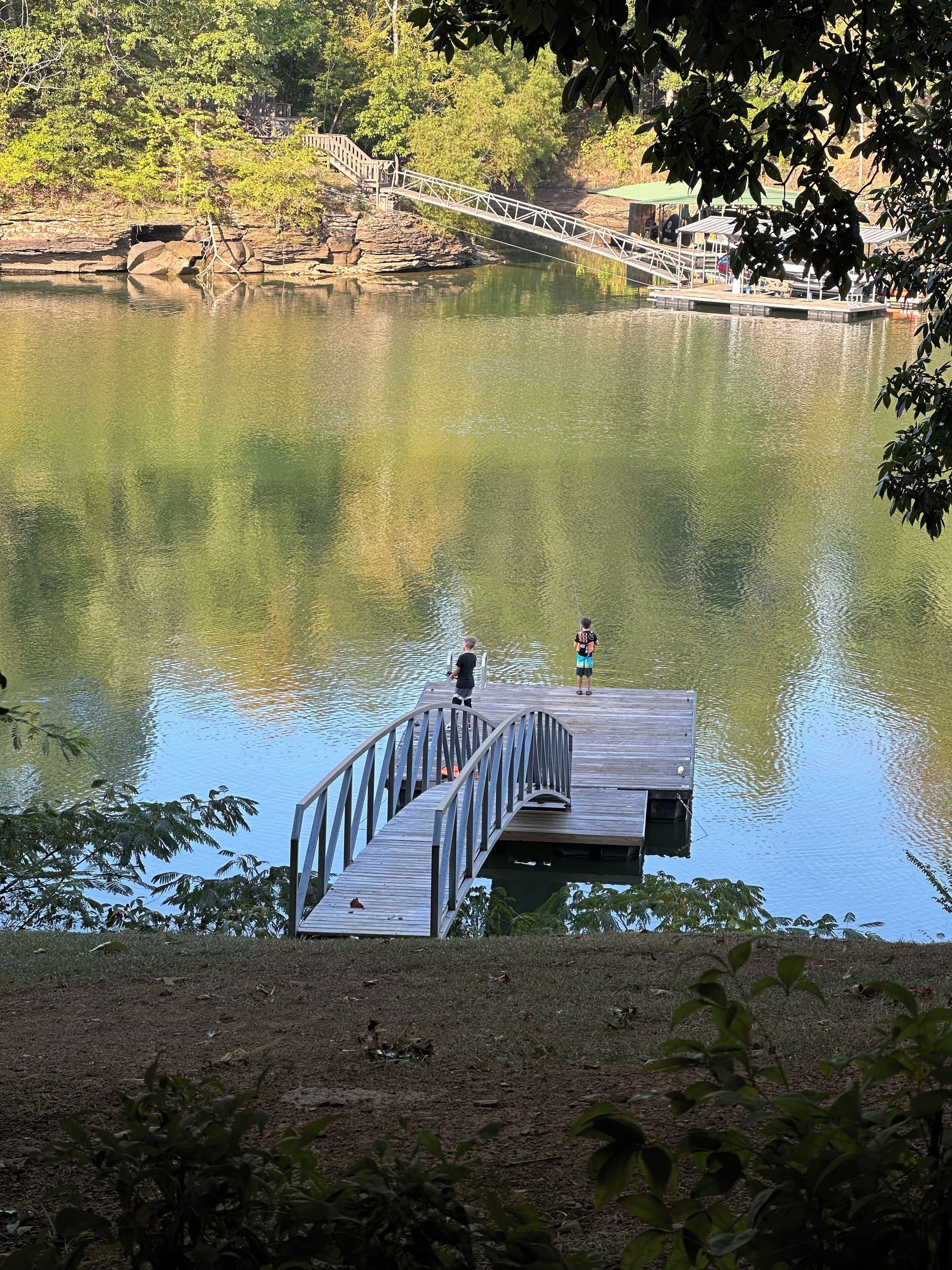 A wooden dock extending into a calm lake, surrounded by trees and a stone bridge in the background.