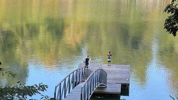 A wooden dock extending into a calm lake, surrounded by trees and a stone bridge in the background.