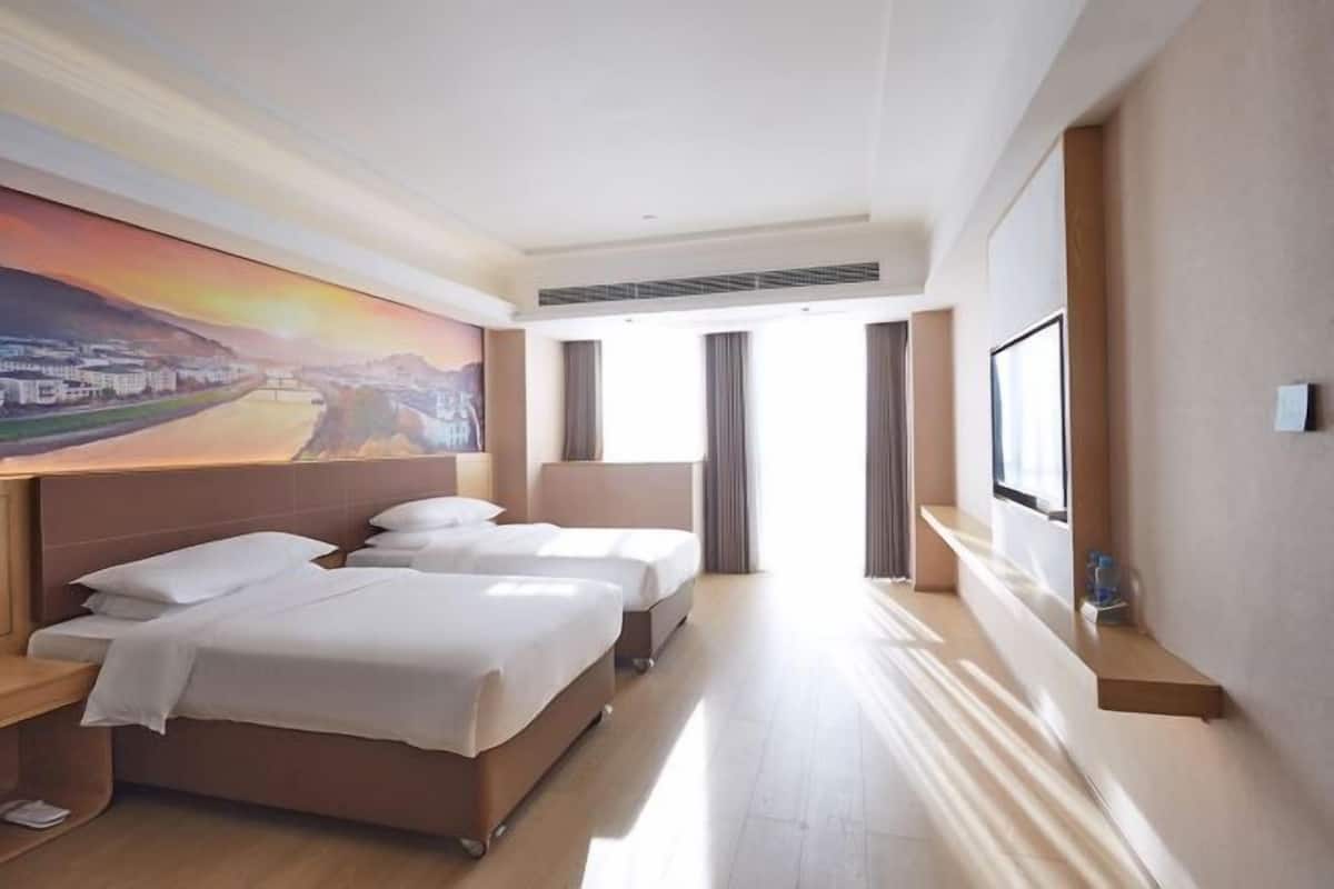A hotel room with two beds, a large painting of a cityscape, a window with curtains, and a built-in shelf.