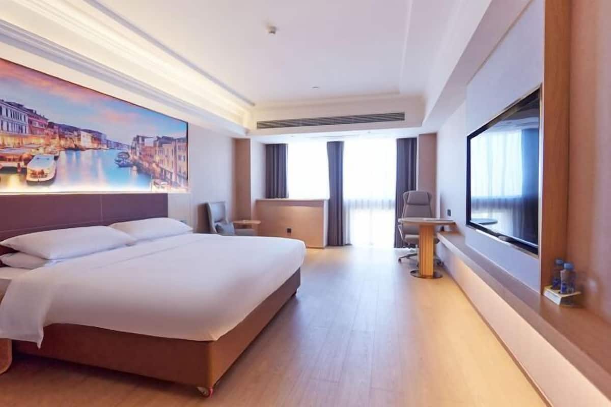 A hotel room with a large bed, a desk, a chair, a TV, and a painting of a cityscape.