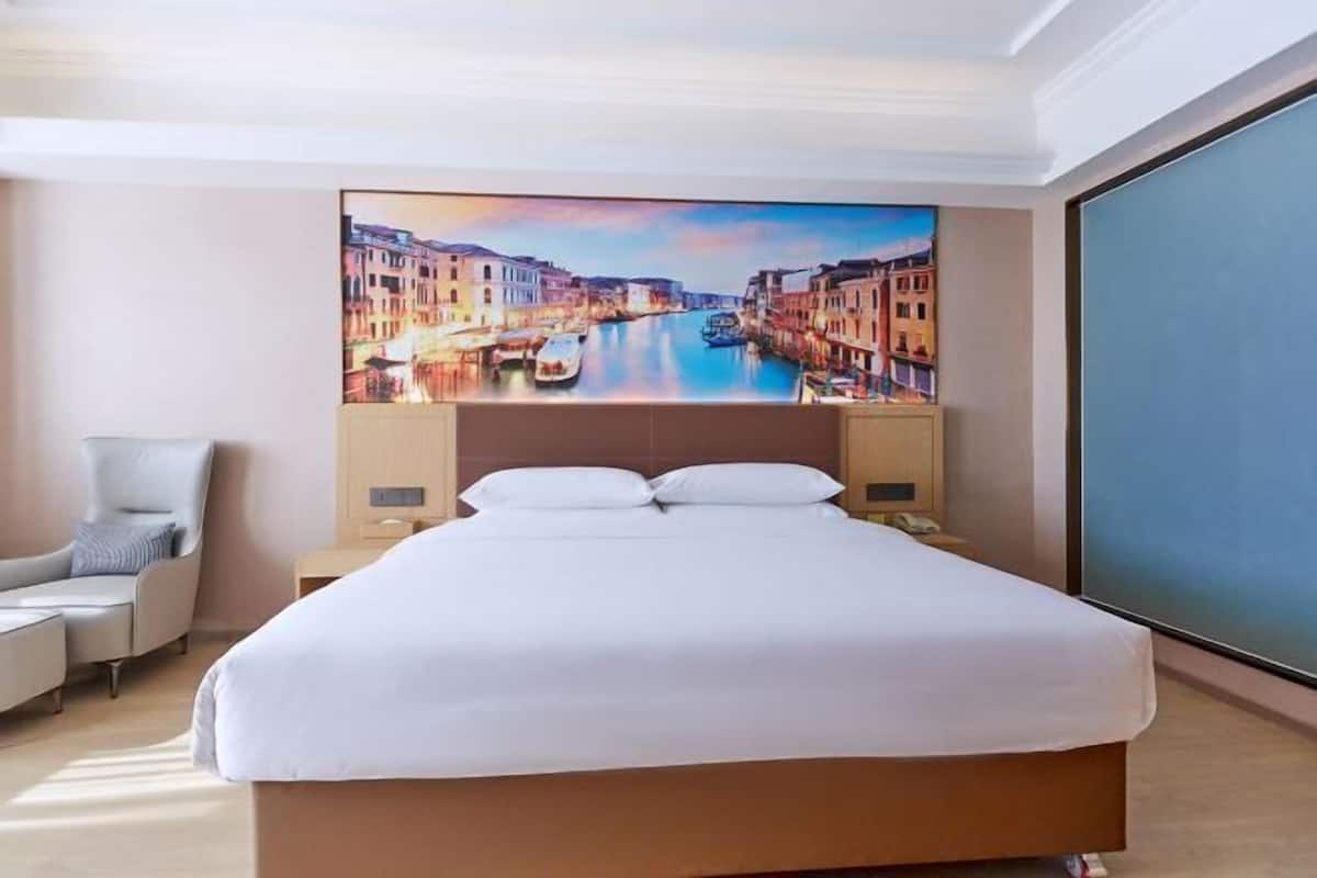 A hotel room with a large bed, a chair, a nightstand, and a painting of a canal scene on the wall.