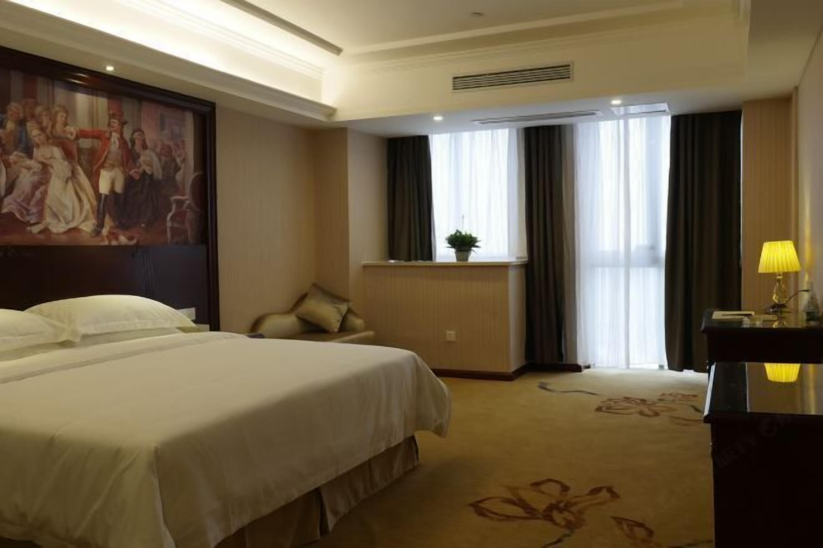 Una habitación de hotel con una cama grande, un escritorio con lámpara, una ventana con cortinas y un cuadro en la pared.