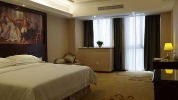 Una habitación de hotel con una cama grande, un escritorio con lámpara, una ventana con cortinas y un cuadro en la pared.