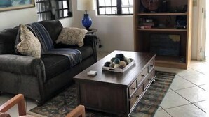 A living room with a dark-colored sofa, a wooden coffee table with decorative items, a blue lamp, and a bookshelf. - Misty's Point of View (St Thomas)