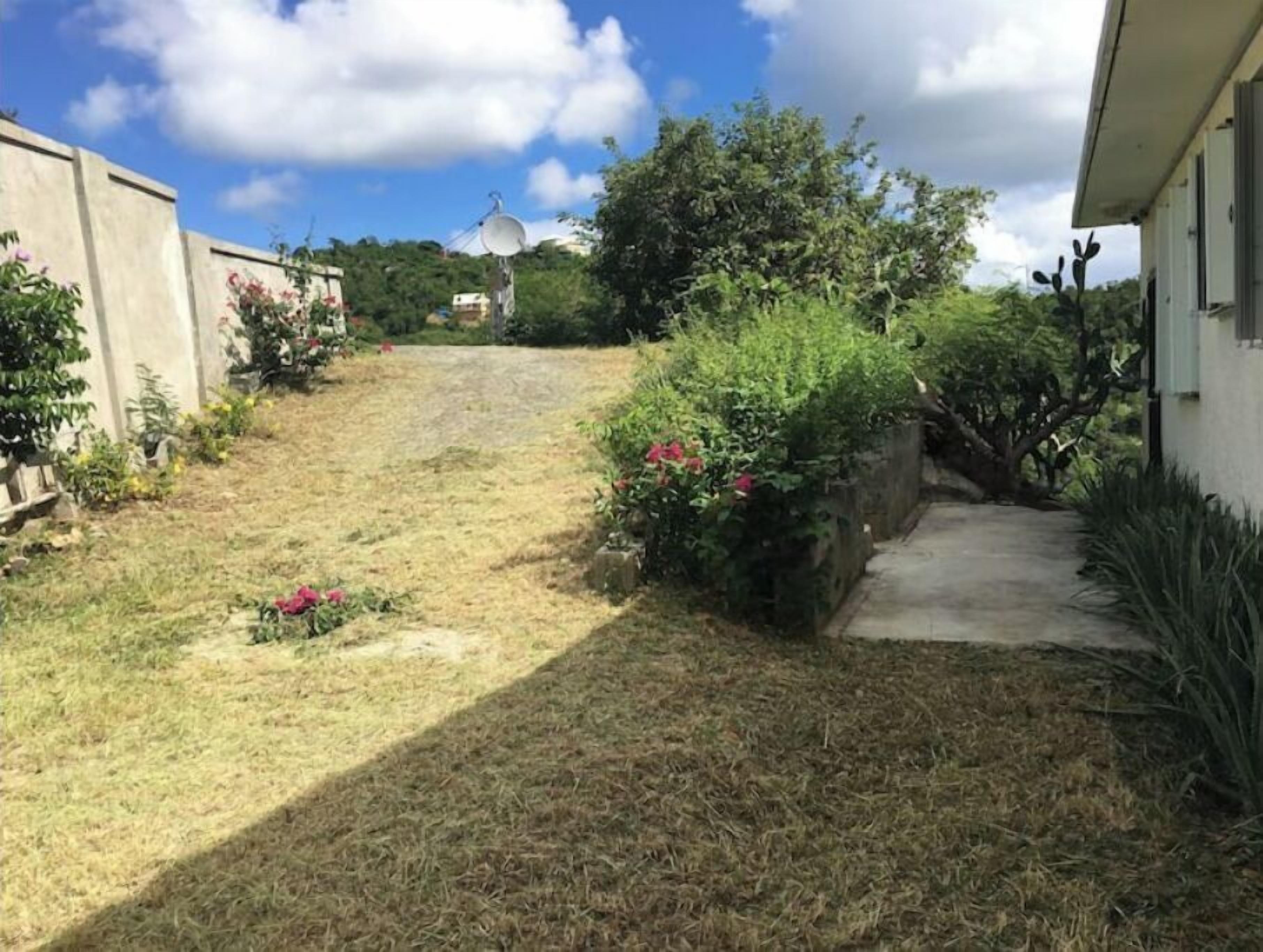 Un étroit sentier mène à une maison surmontée d’une antenne parabolique, le tout étant entouré de verdure et baigné d’un ciel dégagé.
