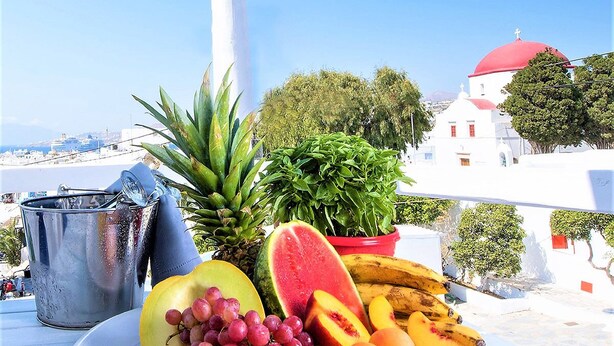 Una mesa con una bandeja de frutas, un cubo metálico y un edificio blanco con cúpulas rojas al fondo.