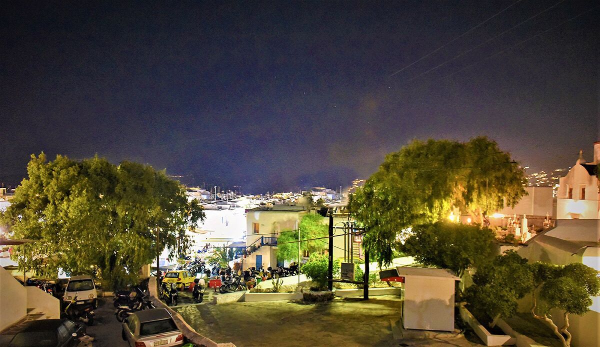 A nighttime view of a coastal town with parked vehicles and trees in the foreground.