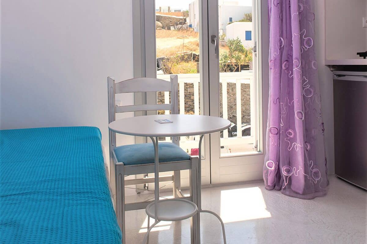A room with a bed, a table, and a chair. There is a window with a view of buildings and a curtain.