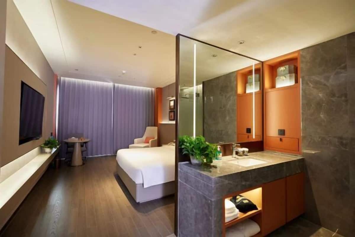 Moderni hotellihuone, jossa on suuri sänky, peilillä varustettu kylpyhuone ja pieni pöytä, jonka päällä on kasvi.