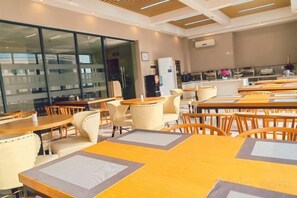 Een restaurantgedeelte in een hotel met tafels en stoelen, een keuken op de achtergrond, en een groot raam met zonwering.