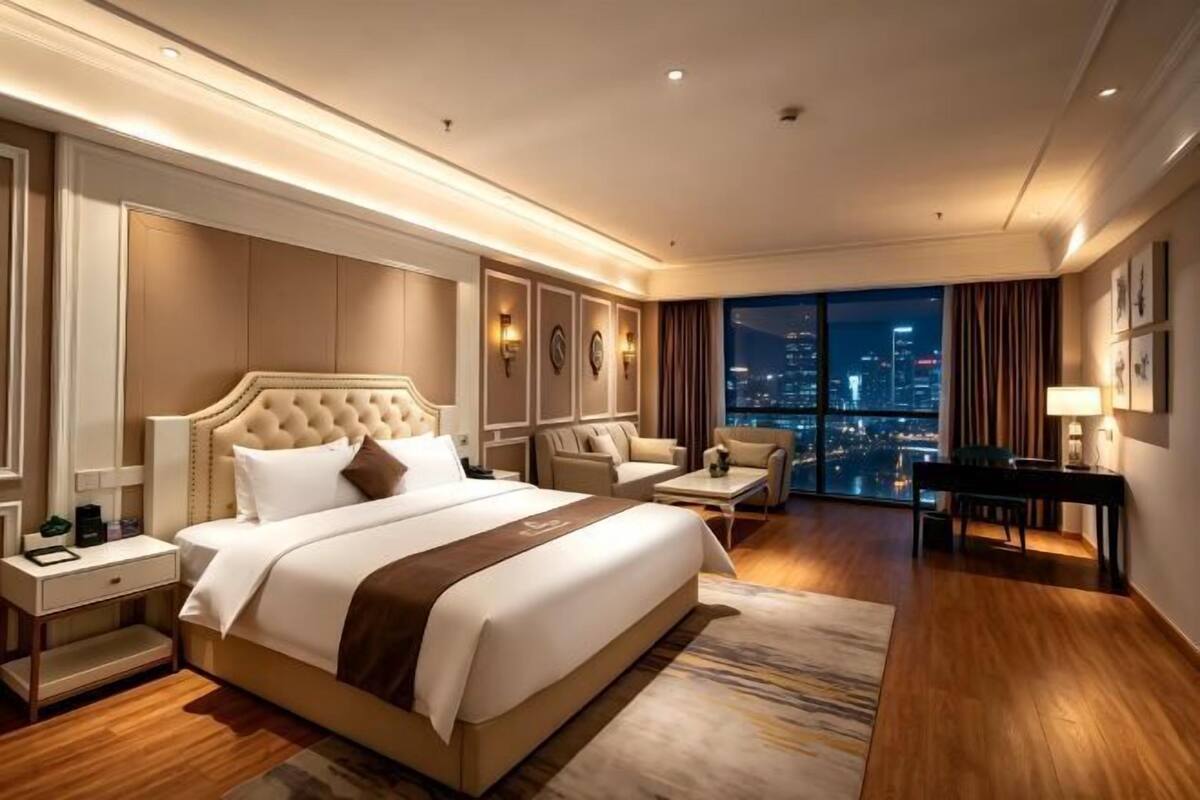 Une chambre d’hôtel avec un grand lit, un coin salon comprenant un fauteuil et une table, une table de chevet et une vue sur la ville la nuit.