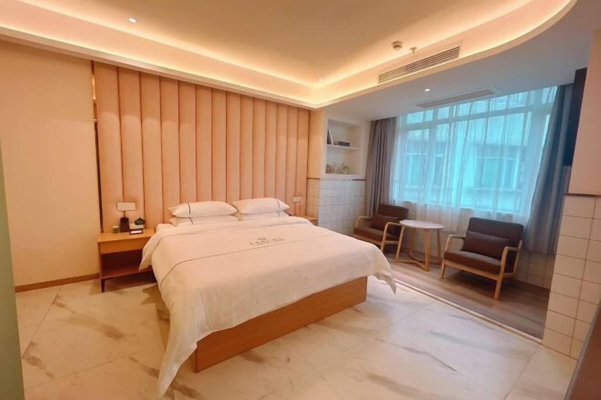 Une chambre d’hôtel moderne dotée d’un grand lit, de tables de chevet, d’un petit coin salon et d’une grande fenêtre avec des rideaux.