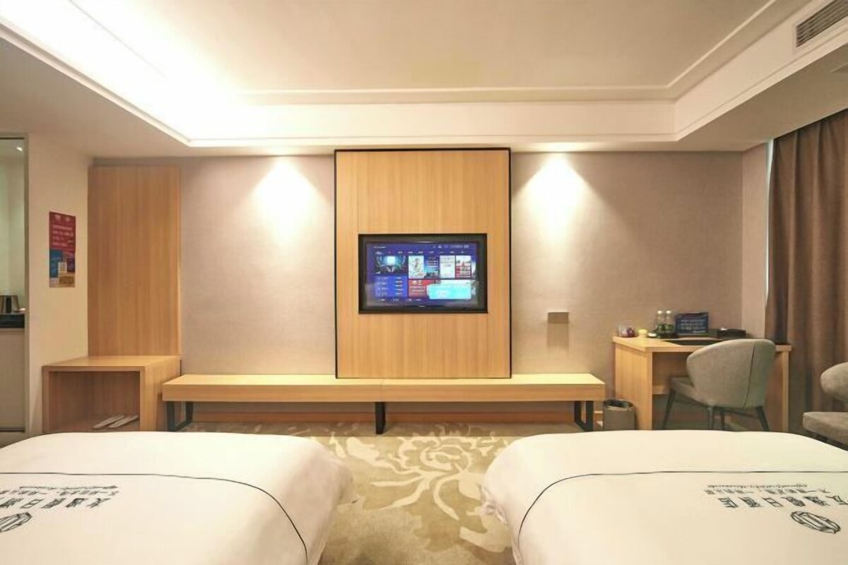 A hotel room with two beds, a desk, a chair, a TV mounted on the wall, and a small table with a lamp.