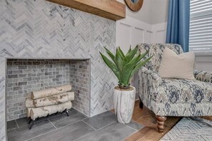 Uma sala de estar com parede em padrão herringbone, lareira com lenha, poltrona estampada e planta em vaso.