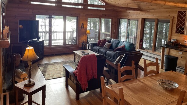 Ein gemütliches Blockhaus-Interieur mit Holzbalkendecke, großen Fenstern, einem Ledersofa, einem Fernseher und einem Essbereich.