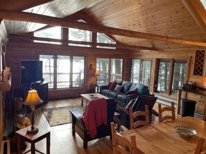 A cozy cabin interior with a wooden ceiling, large windows, a leather sofa, a TV, and a dining area. - Lakefront Hemlock house on 3 acres, 5-Bdrm retreat on private lake, near casino (Minocqua)
