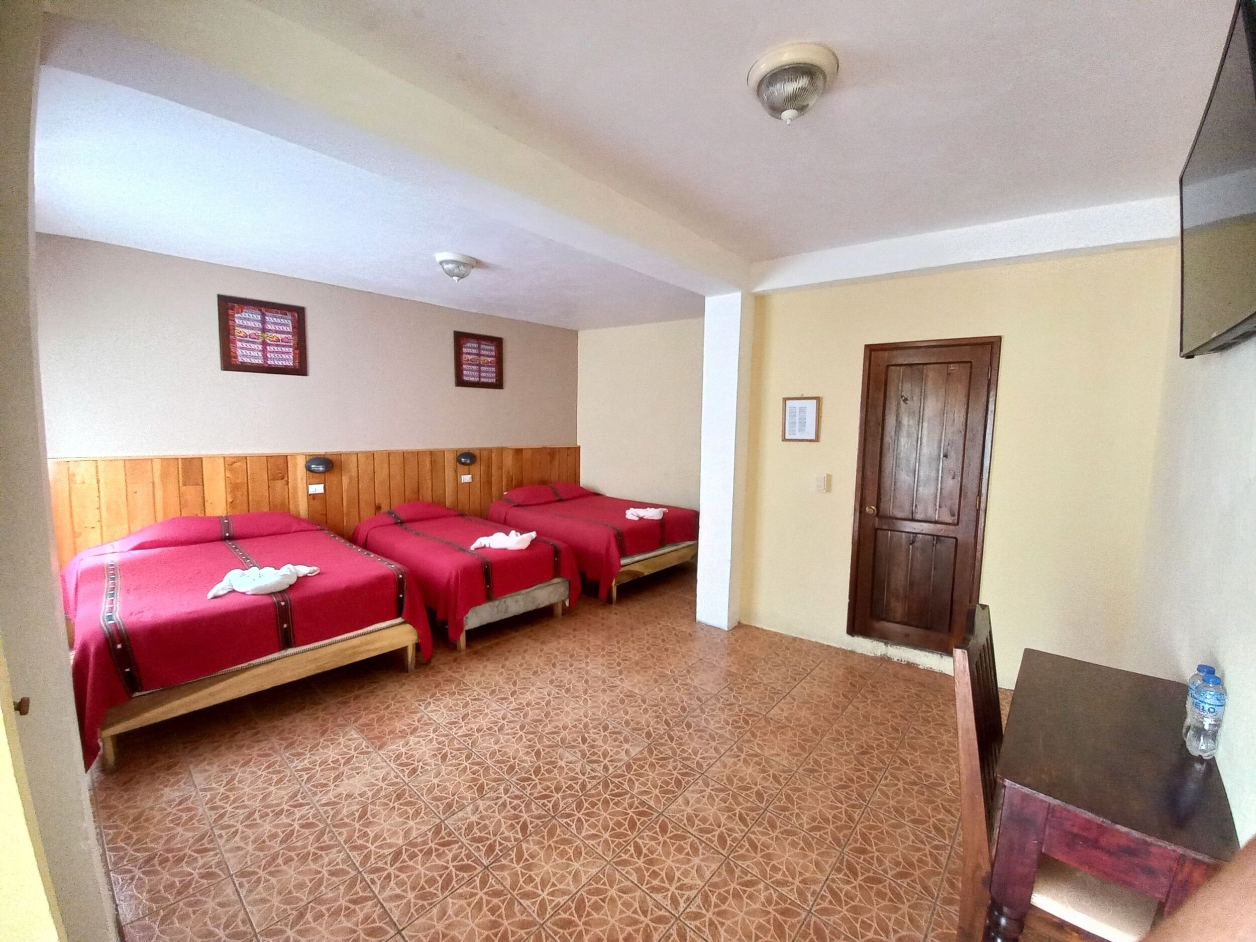 Una habitación con cuatro camas, cabecera de madera, piso de baldosas, puerta de madera y ventilador de techo.