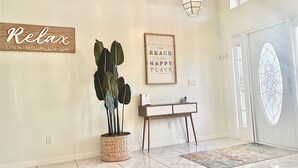 A hallway with a wooden sign saying "Relax Unwind & Enjoy," a framed quote on the wall, a potted plant, a console table, and a decorative glass door. - Stunning Sunsets-Heated Pool and Spa-Golf Course Views (Cape Coral)