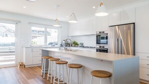 A modern kitchen with white cabinetry, stainless steel appliances, and wooden bar stools. - Salt and Light Mollymook (Mollymook Beach)