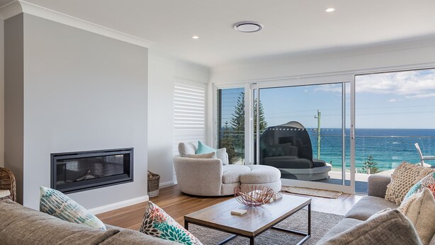 A modern living room with a fireplace, a sofa, a coffee table, and a view of the ocean through large glass doors. - Salt and Light Mollymook (Mollymook Beach)