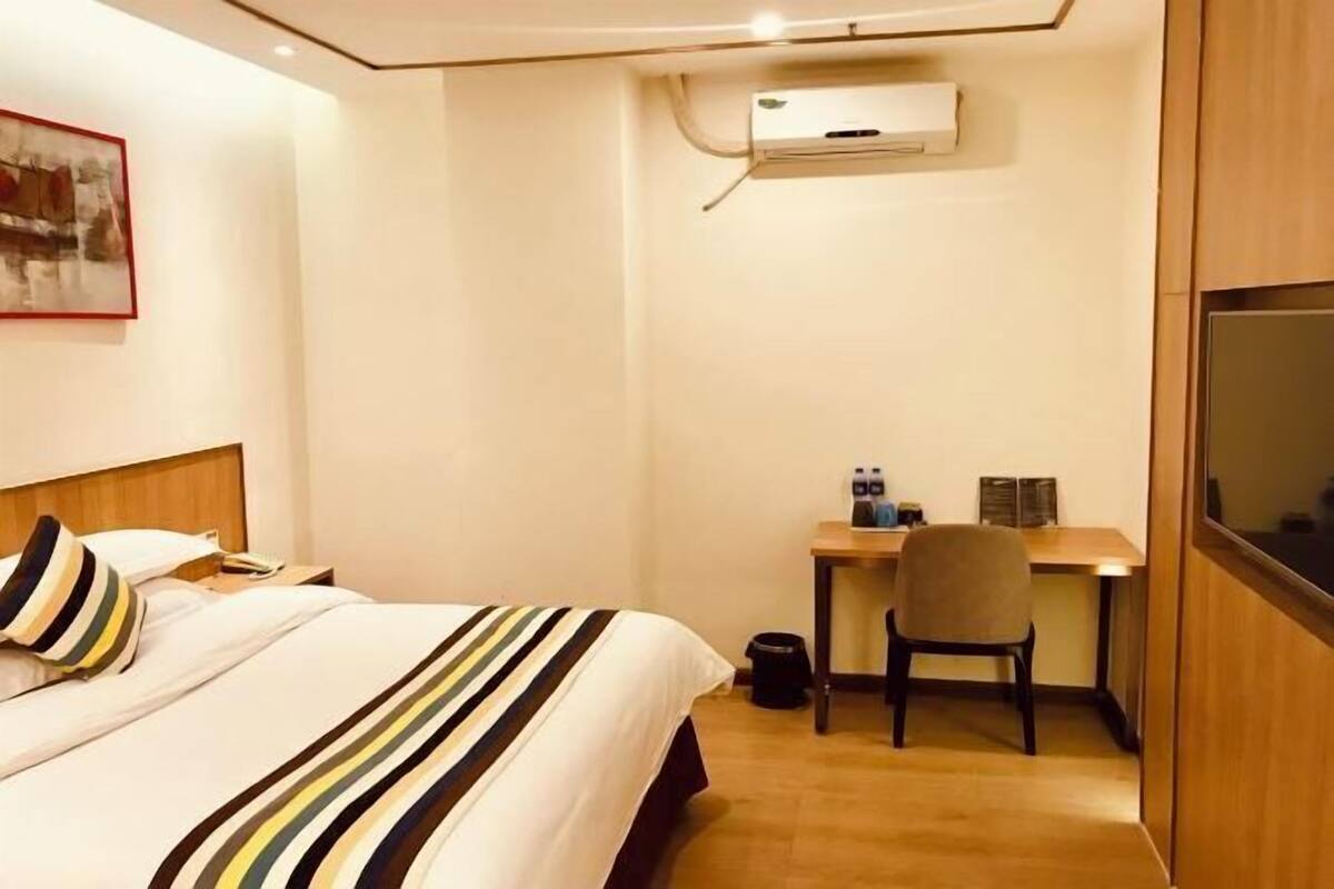 A hotel room with a bed, a desk, a chair, and an air conditioning unit.