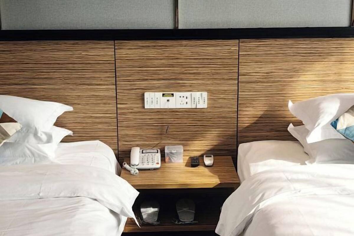 A hotel room with a wooden headboard, a bedside table with a phone, a small container, and a remote control.