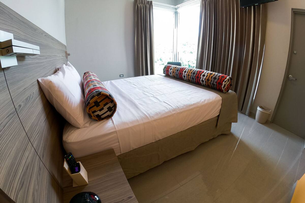 A modern bedroom with a wooden headboard, a large bed with a patterned pillow, a bedside table with a remote control, and a window with curtains.