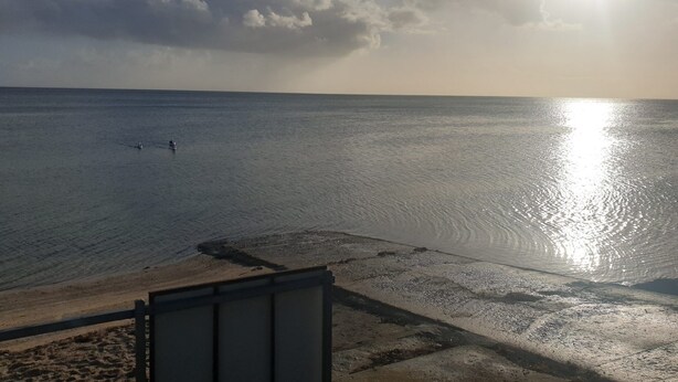 Una playa con cielo despejado, el sol reflejado en el agua y algunas personas a lo lejos.