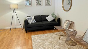 Una sala de estar con un sofá negro, dos almohadones estampados, una lámpara de pie, un espejo redondo y tres láminas botánicas enmarcadas en la pared.