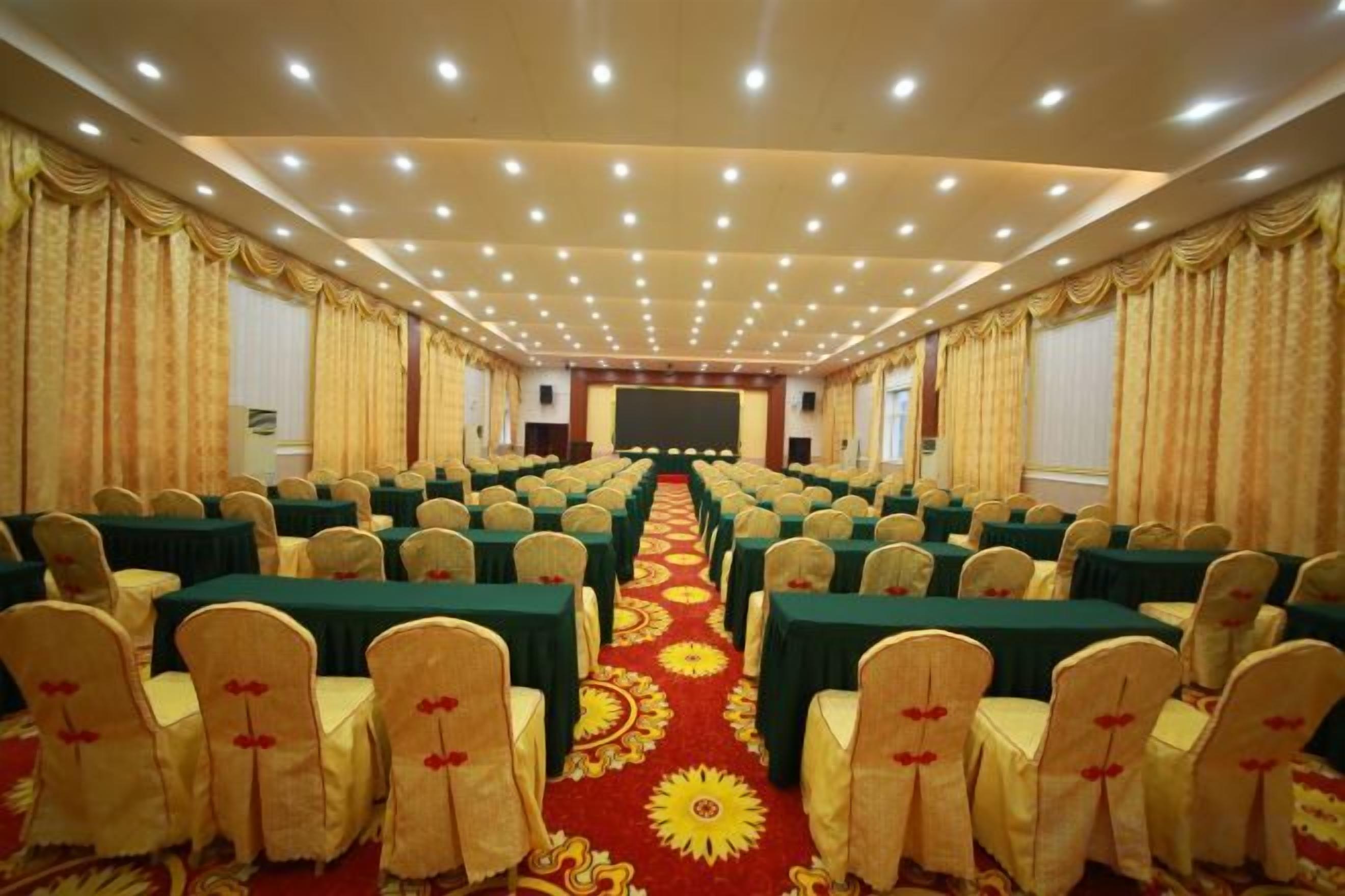 Un salón de conferencias con filas de mesas y sillas, piso alfombrado y cortinas en las ventanas.