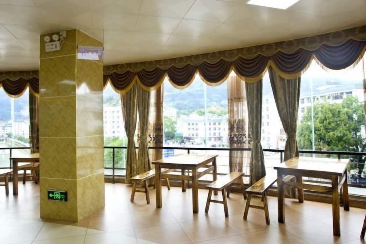 A dining area with wooden tables and benches, large windows with curtains, and a view of buildings and greenery.