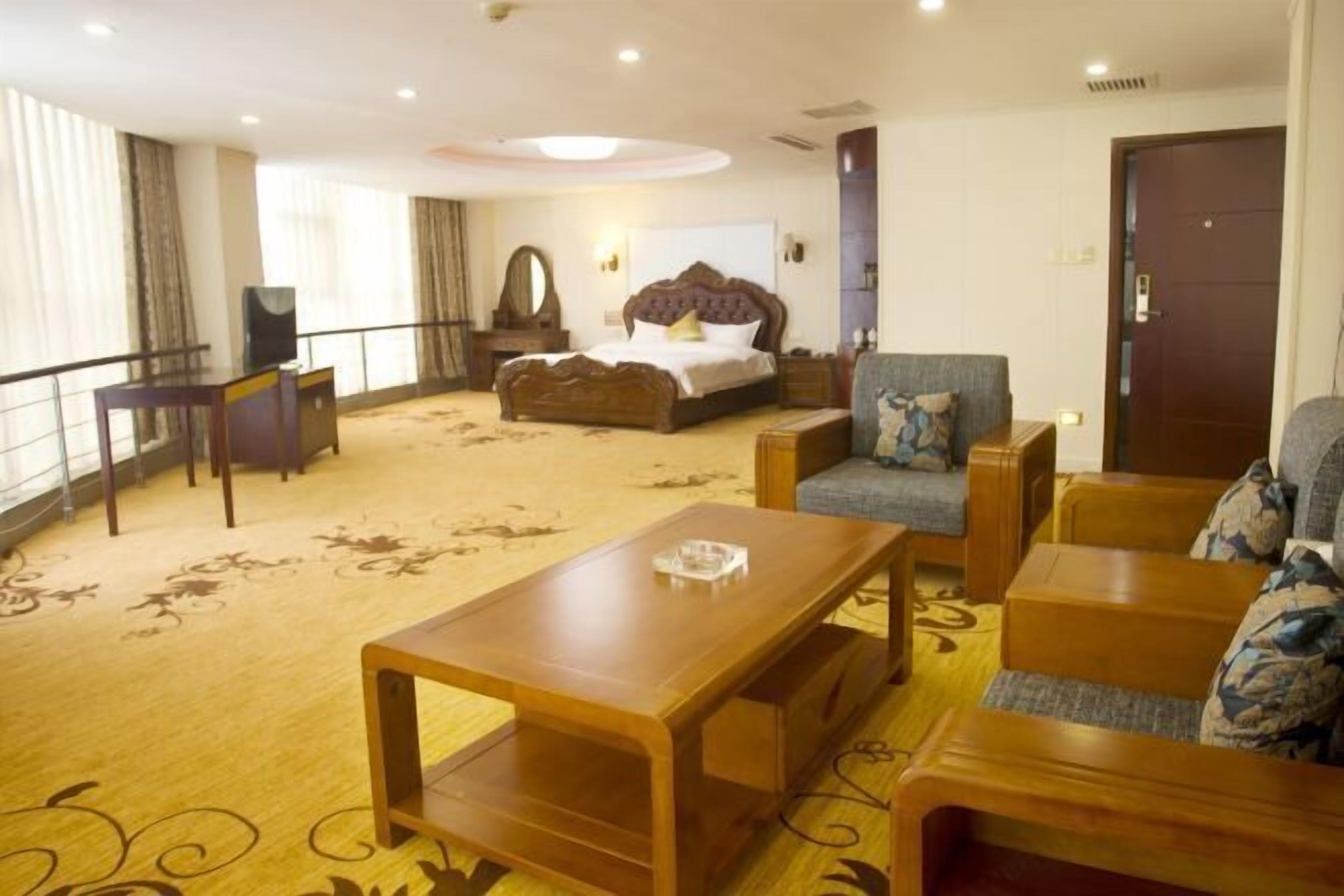 A hotel room with a bed, a sofa, a wooden coffee table, a TV, and a large window with curtains.