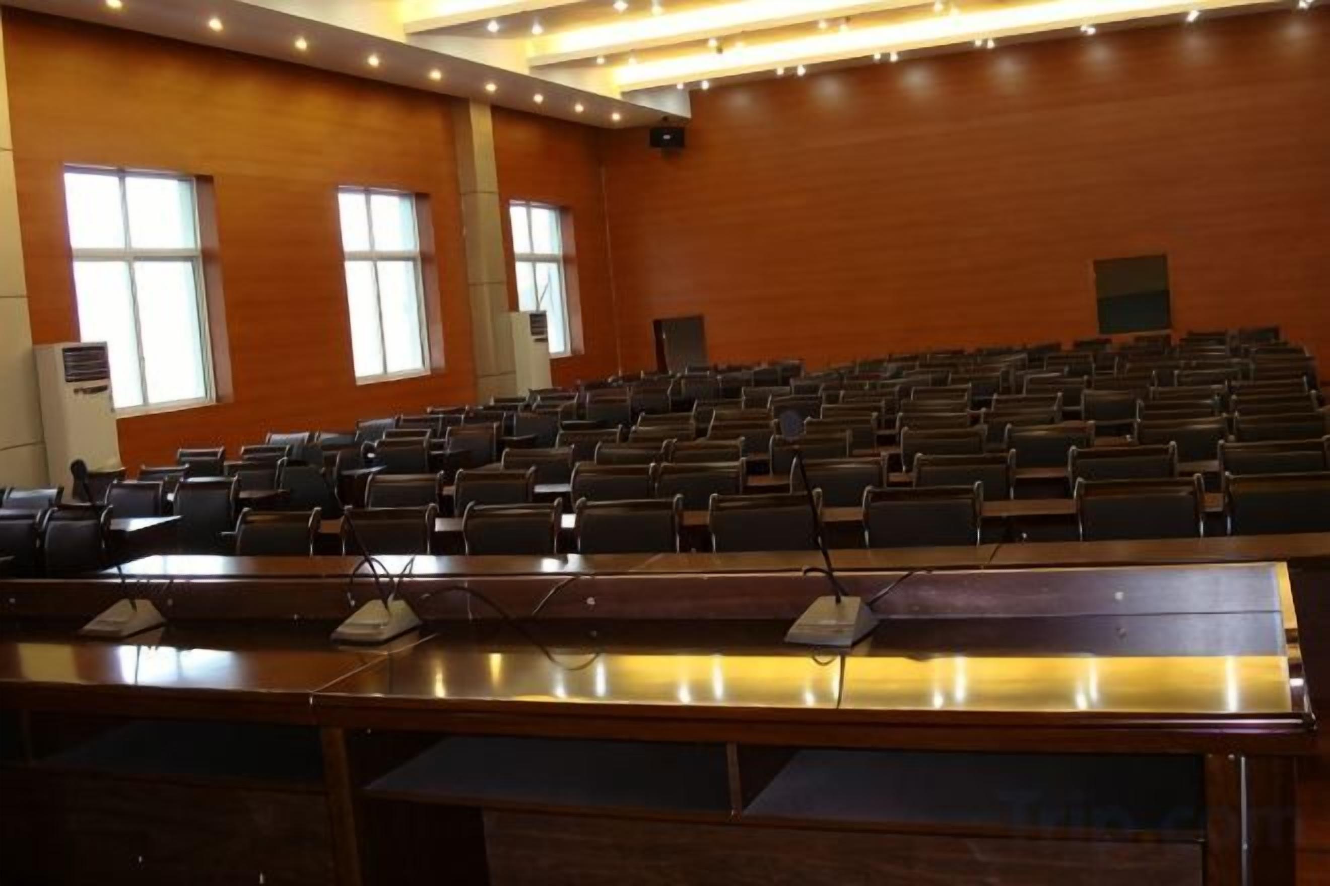 A conference room with rows of chairs, a long table with microphones, and large windows.