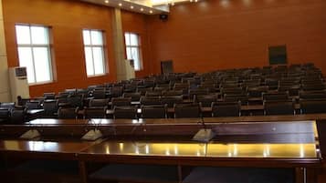 A conference room with rows of chairs, a long table with microphones, and large windows.