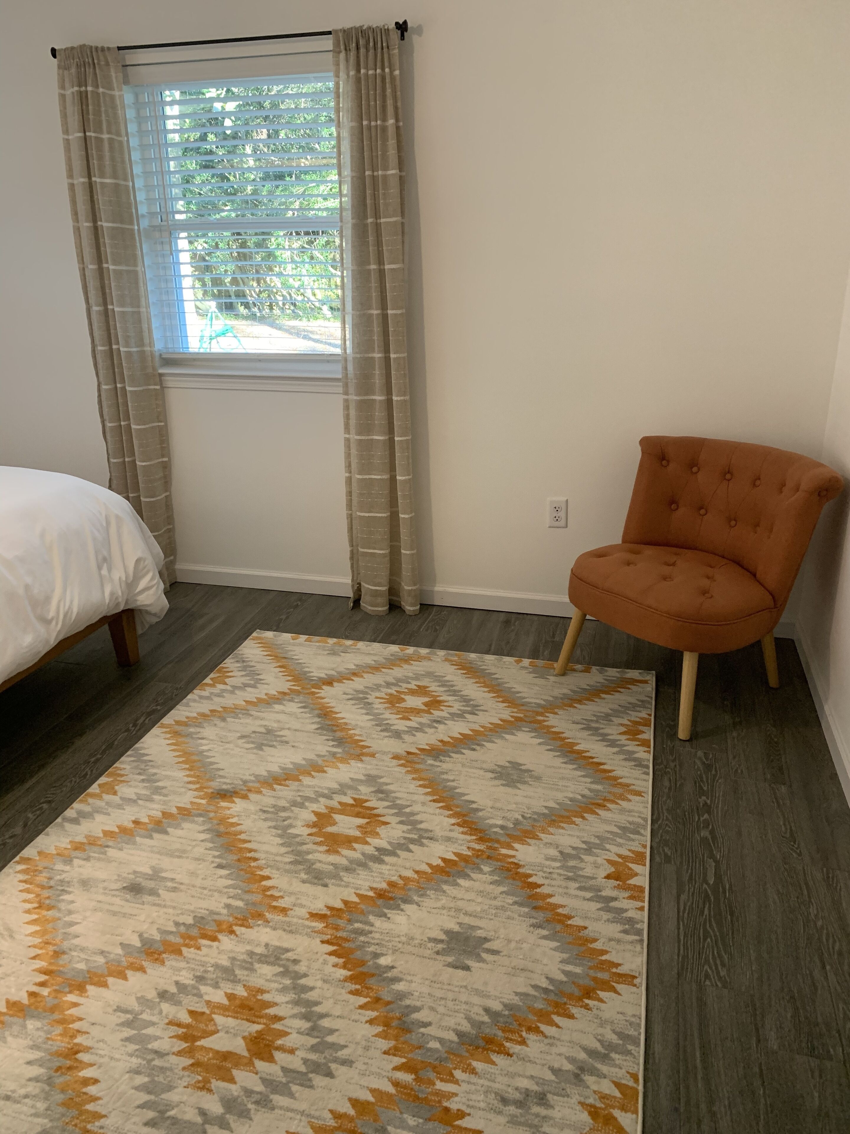 Un dormitorio con cama, alfombra estampada, ventana con cortinas y una silla.