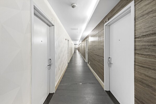 A long, narrow hotel corridor with doors on both sides, a patterned wall on the left, and a wooden wall on the right.